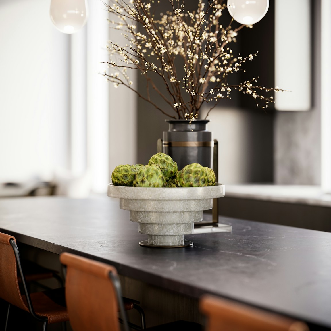 A modern kitchen features a dark countertop with a stone bowl holding green objects. Behind it, a vase with branches of small white flowers adds a delicate touch. Leather chairs are lined up along the counter. The space is sleek and contemporary.